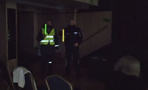 Policjant stoi obok ubranego w kamizelkę odblaskową mężczyzny. Zgaszone światła w pomieszczeniu. Obaj trzymają w ręce odblaskową opaskę.