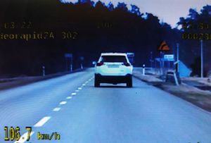 Obraz widziany okiem kamery z radiowozu. Na jezdni biały samochód typu suv widziany z tyłu. W lewym dolnym narożniku kadru wyświetlono prędkość 106.7 km/h
