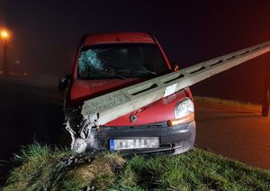 Pora nocna,złamany betonowy słup oświetleniowy przygniata pokrywę silnika czerwonego renaulta kangoo, pojazd ma uszkodzoną przednią szybę , w tle widoczna  stojąca latarnia.
