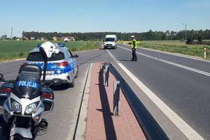 Policyjny motocykl i radiowóz, stoją na pasie do kontroli drogowej. Za nimi na jezdni policjant daje znak do zatrzymania dla nadjeżdżającego białego dostawczego pojazdu. W tle obszar rolniczy i drzewa.