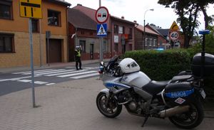 Na pierwszym planie policyjny motocykl, za nim na przejściu dla pieszych stoi policjant ruchu drogowego i zatrzymuje ruch pojazdów.