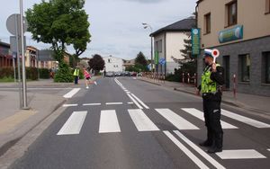 Policjant drogówki zatrzymuje ruch trzymając tarczę sygnałową. W tle biegnący uczestnik zawodów oraz drugi policjant zatrzymujący z ruch z przeciwnego kierunku.