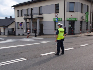Policjant ruchu drogowego stoi na jezdni wstrzymując ruch pojazdów. Przed nim wybiegający z drogi podporządkowanej uczestnik zawodów.