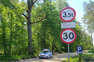Droga leśna w tle radiowóz. Po lewej stronie drogi znaki informujące o obowiązującej prędkości 30 km/h oraz obowiązującym tonażu pojazdów