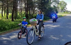 Kobieta na rowerze. Obok niej dziecko na małym rowerku z kasku i kamizelce odblaskowej. Za policjant na motocyklu z włączonymi sygnałami świetlnymi.