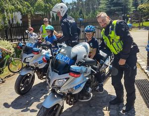 Dwa policyjne motocykle. Obok nich policjanci. Jeden w hełmie ochronnym. Na motocyklach siedzą dzieci i pozują do zdjęcia.