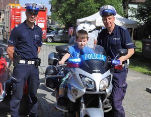 Policjanci ruchu drogowego pozują do zdjęcia. Pomiędzy nimi stoi zaparkowany motocykl z napisem na owiewce POLICJA. Na motocyklu siedzi mały chłopiec w niebieskiej koszulce. Prawa rękę trzyma na kierownicy.