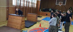 Sala gimnastyczna. Instruktor demonstruje jeden ze sposobów pokonywania skrzyni gimnastycznej. Obok stoi grupa młodzieży.