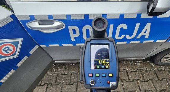 Miernik prędkości z zapisanym odczytem prędkości 110 km/ h, tło stanowi boczna część radiowozu z napisem policja.