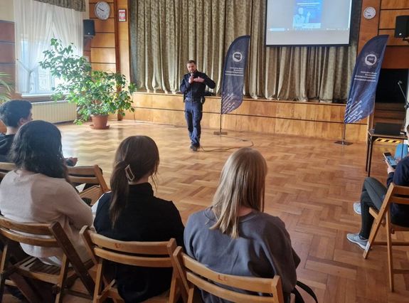 uczniowie siedzą w auli Zespołu Szkół w Olesnie, na środku stoi policjant, który w prawej dłoni trzyma mikrofon.