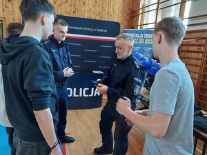 Zdjęcie w kolorze, przedstawia młodych mężczyzn stojących z policjantem, który trzyma w dłoniach opaskę zaciskową do tamowania krwotoków typu &quot; Staza&quot;. W tle granatowa ściana z płótna z napisem Komenda Powiatowa Policji w Olesnie, oraz granatowy pulpit z napisem Policja.