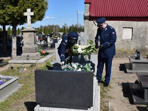 Zdjęcie w kolorze przedstawia cmentarz, dwaj policjanci w mundurach galowych w kolorze granatowym pochylają się nad grobem. Policjant po lewej stronie trzyma w rękach kwiaty przewiązane biało czerwoną wstęga, drugi policjant trzyma w ręku znicz. W tle inne mogiły i fragment błękitnego nieba.