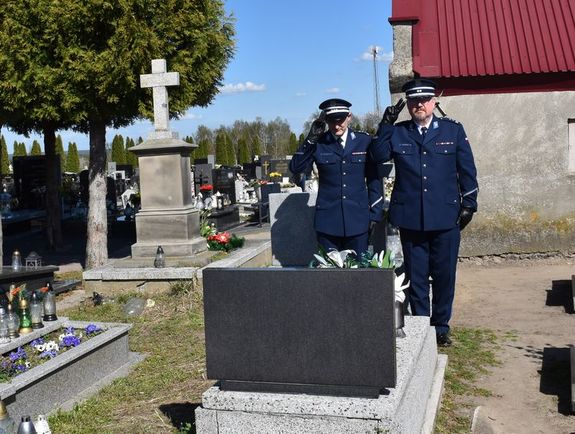 Zdjęcie w kolorze, przedstawia dwóch policjantów w wyjściowych granatowych mundurach, stoją na baczność i w geście oddania honoru trzymają prawe złączone palce przy skroniach. W tle groby i fragment błękitnego nieba.