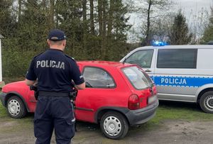 Zdjęcie w kolorze przedstawia policjanta stojącego przed czerwonym oplem, zaraz za nim stoi policyjny radiowóz z włączonymi niebieskimi światłami, w tle drzewa i jezdnia.