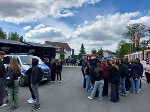 Zdjecie w kolorze przedstawia grupę młodzieży około 100 osób stojących przed czarnym pojazdem cięzarowym. Po lewej stronie dwa policyjne pojazdy. W tle niebo z białymi obłokami.