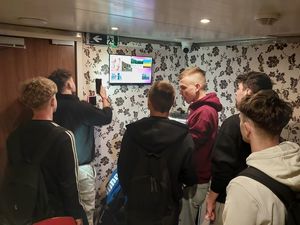 Zdjecie w kolorze przedstawia grupę młodych mężczyzn wewnątrz pokoju, w którym spoglądają na monitor zawieszony na ścianie.