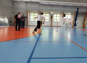 Zdjęcie w kolorze przedstawia młodego meżczyznę który biega pomiędzy tyczkami wyznaczającymi tak zwaną kopertę. Obok po lewej stronie stoją policjant i policjantka.