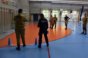 Zdjęcie w kolorze przedstawia mężczyznę w piaskowym mundurze z napisem ochotnicza straż pożarna, który trzyma w rekach piłke 3 kilogramową. Obok po prawej stronie stoi policjantka kilka metrów dalej stoją młodzi meżczyźni w podobnych mundurach.