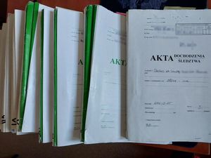 Zdjęcie w kolorze przedstawia stos ułożonych częściowo jedna na drugiej teczce w kolorze szarym z napisem Akta dochodzenia/ śledztwa.