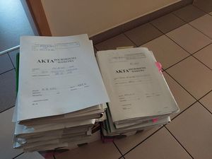 Zdjęcie w kolorze przedstawia, teczki w kolorze szarym z napisem Akta dochodzenia/ Śledztwa ułożone jedna na drugiej w dwóch stertach na wykafelkowanej podłodze.