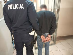 Zdjęcie w kolorze przedstawia policjanta który trzyma za rękę zatrzymanego. Obaj stoją obok drzwi - kraty w pomieszczeniu dla osób zatrzymanych.