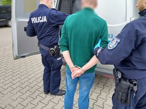 Zdjęcie w kolorze przedstawia policjantów którzy stoją z zatrzymanym przy tylnych drzwiach radiowozu. Mężczyzna jest trzymany przez policjanta z prawej strony, ma kajdanki założone na ręce trzymane z tyłu.