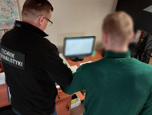 Zdjęcie w kolorze przedstawia technika kryminalistyki, który trzyma lewą dłoń zatrzymanego mężczyzny na urządzeniu do skanowania linii papilarnych.