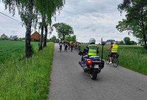 Zdjęcie w kolorze przedstawia policjanta na motocyklu jadącego na końcu peletonu. W tle drzewa i chmury oraz początek zabudowań wiejskich.