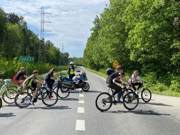 Zdjęcie w kolorze przedstawia rowerzystów przejeżdżających przed jezdnię do lasu, tuż za nimi policjant wstrzymujący ruch.