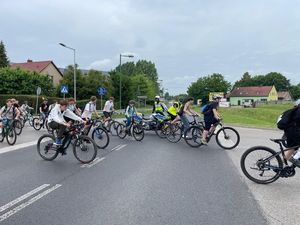 Zdjęcie w kolorze przedstawia rowerzystów przejeżdżających przez skrzyżowanie w mieście, tuz za nimi policjant wstrzymujący ruch.