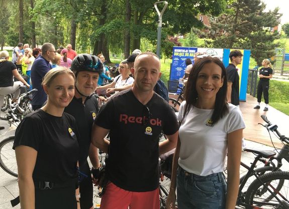 Zdjęcie w kolorze przedstawia dwie kobiety stojące po prawej i lewej stronie mężczyzny w tle stojące rowery i inni uczestnicy.