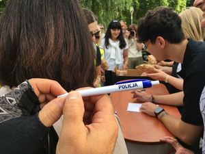 Zdjęcie w kolorze przedstawia na pierwszym planie  trzymany w dłoniach długopis z napisem policja w tle uczestnicy wydarzenia.