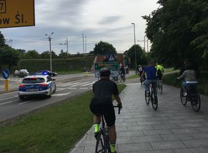 Zdjęcie w kolorze, przedstawia rowerzystów jadących deptakiem oraz jezdnią , po lewej stronie radiowóz na jezdni - zabezpiecza peleton.