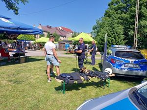 zdjęcie w kolorze przedstawia pojazdy policyjne zaparkowane na trawniku przed nimi stoja policjanci z psem służbowym obok mężczyzna a w tle inne osoby - uczestniczace w pikniku.