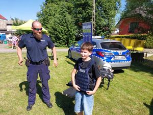 Zdjęcie w kolorze przedstawia chłopca w wieku około 8 lat który ma załozona kamizelkę balistyczna z napisem policja. Obok niego stoi uśmiechnięty policjant w okularach przeciwsłonecznych z brodą.