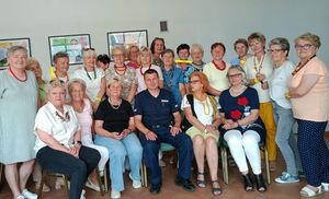 Zdjęcie w kolorze przedstawia seniorów pozujących do zdjęcia osoby z tyłu stoją a osoby w pierwszym szeregu siedzą na krzesłach, po środku policjant.