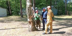 Plac apelowy obozowiska harcerskiego w Kucobach. Policjant i strażak z Państwowej Straży Pożarnej rozmawiają z jedną z opiekunek. W tle pomiędzy drzewami rozbite namioty.
