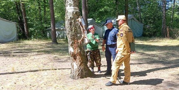 Plac apelowy obozowiska harcerskiego w Kucobach. Policjant i strażak z Państwowej Straży Pożarnej rozmawiają z jedną z opiekunek. W tle pomiędzy drzewami rozbite namioty.
