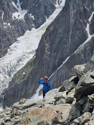 Alpinista wspina się skalnym podejściem. W tle skaliste zbocze góry w żlebach leży śnieg.