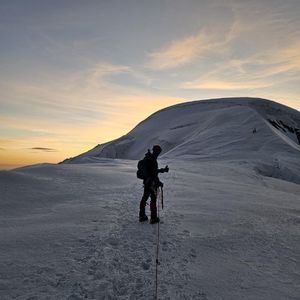Alpinista w kasku ochronnym i z plecakiem na plecach stoi na ośnieżonym stoku. w tle pasmo górskie.