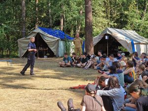 Zdjęcie w kolorze, przedstawia uczestników obozu - dzieci i młodzież siedzących na leśnej polanie. Na środku przed nimi stoi policjantka. W tle drzewa i namioty.