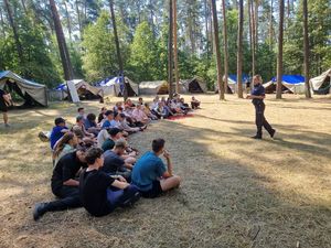 Zdjęcie w kolorze, przedstawia uczestników obozu po lewej stronie - siedzących na leśnej polanie, tuż za nimi namioty i drzewa . Po prawej stronie  stoi policjantka. W tle drzewa i namioty.