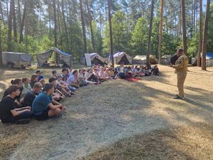 Zdjęcie w kolorze, przedstawia uczestników obozu po lewej stronie - siedzących na leśnej polanie, tuż za nimi namioty i drzewa . Po prawej stronie  stoi funkcjonariusz Państwowej Straży Pożarnej. W tle drzewa i namioty.