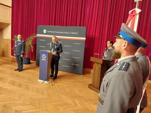 Zdjęcie w kolorze, przedstawia Komendanta Powiatowego Policji w Oleśnie podczas przemówienia w sali Miejskiego Domu Kultury w Oleśnie, z lewej strony stoi Zastępca Komendanta Wojewódzkiego insp. Krzysztof Buchała, za nimi ścianka z tkaniny z napisem Komenda Powiatowa Policji w Oleśnie. Po prawej stronie fragment Pocztu Sztandarowego oleskiej policji.