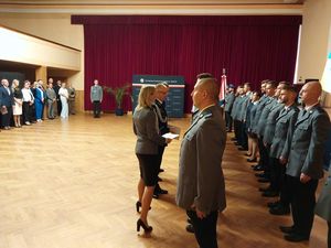 Zdjęcie w kolorze, przedstawia Policjantów w dwóch szeregach stojących po prawej stronie, na środku Zastępca Komendanta Wojewódzkiego insp. Krzysztof Buchała w asyście policjantki wręcza akty mianowania na wyższe stopnie policyjne.