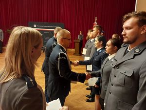 Zdjęcie w kolorze, przedstawia Policjantów w dwóch szeregach stojących po prawej stronie, na środku Zastępca Komendanta Wojewódzkiego insp. Krzysztof Buchała w asyście policjantki wręcza akty mianowania na wyższe stopnie policyjne.