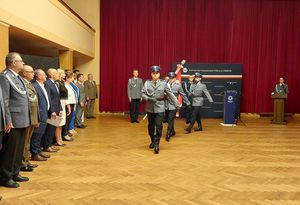 Zdjęcie w kolorze przedstawia Policjantów ze Sztandarem Komendy Powiatowej Policji w Oleśnie podczas wymarszu z sali, za nimi ściana z granatowego płótna z napisem Komenda Powiatowa Policji w Oleśnie. Po lewej stronie stoją Policjanci oraz przedstawiciele instytucji i władz lokalnych.