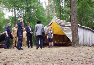 Zdjęcie w kolorze, przedstawia grupę osób w tym funkcjonariuszy policji i straży pożarnej stojących w grupie na obszarze leśnym, w tle namiot harcerski.