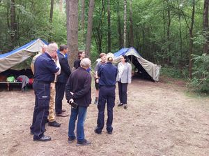 Zdjęcie w kolorze, przedstawia grupę osób w tym funkcjonariuszy policji i straży pożarnej stojących w grupie na obszarze leśnym, w tle namiot harcerski.