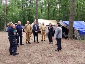 Zdjęcie w kolorze, przedstawia grupę osób w tym funkcjonariuszy policji i straży pożarnej stojących w grupie na obszarze leśnym, w tle namiot harcerski.